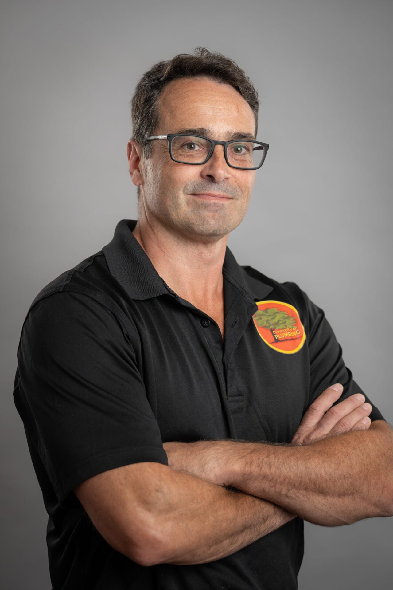 Paul, a confident and professional plumber, stands with arms crossed wearing a black polo shirt with the "Cherry Oak Plumbing" logo, set against a neutral gray background.