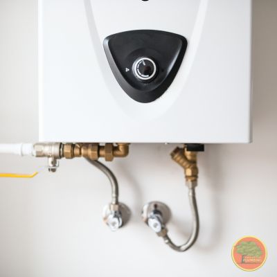 A close-up view of a modern tankless water heater mounted on a wall. The unit features a black central dial for temperature control. Below, connected water lines and valves—both metal and PVC—are neatly installed. The setup appears clean and professionally done. A small circular logo is visible in the bottom right corner.